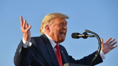 Donald Trump speaking into a microphone against a backdrop of the sky. He is gesticulating with his hands.
